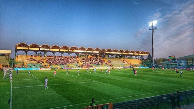 grass, large group of people, built structure, architecture, building exterior, sky, sport, green color, field, blue, lawn, street light, men, outdoors, playing field, grassy, stadium, clear sky, day