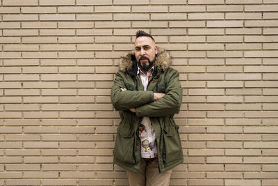 Bearded male with crossed arms leaning on bricked wall, looking camera