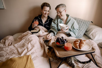 Smiling lesbian couple having breakfast in bed with cat at home