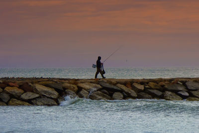 Silhouette man fishing in sea against sky during sunset