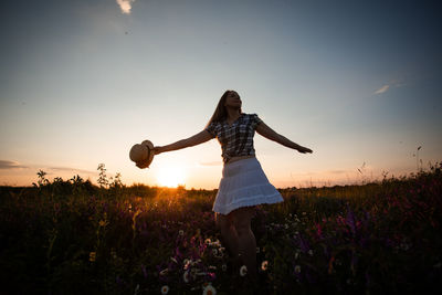 Woman with arms outstretched on field against sky during sunset