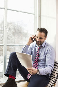 Mid adult businessman reading document while on call by office window