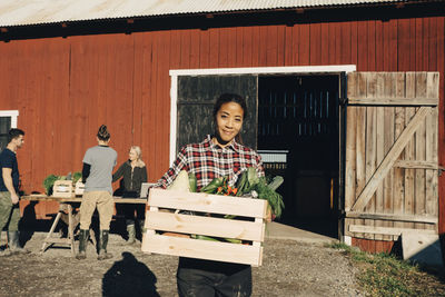 Portrait of happy mature woman carrying crate full of vegetables with barn in background