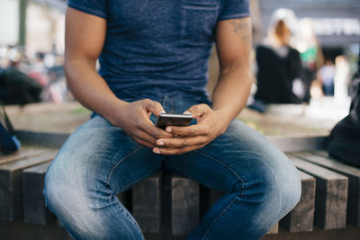 Midsection of young man using smart phone while sitting on bench in city