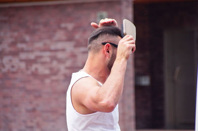 Side view of man brushing hair while standing against wall