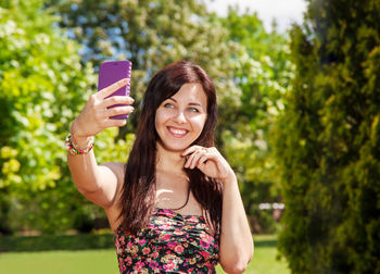 Smiling young woman taking selfie while smart phone while standing against trees in park