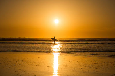 Silhouette surfer standing at beach against sky during sunset