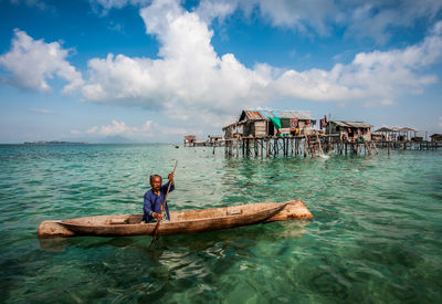 Man oaring boat by stilt houses in sea against cloudy sky