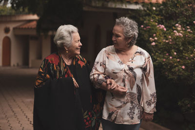 Old ladies wearing casual clothes and having conversation while walking together in summer garden near green bushes of roses on cloudy day