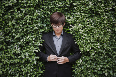 A young asian groom looked down at his hand wearing a suit in front of the tree bush.