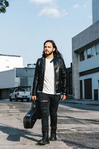 Trendy latin american male guitarist in leather jacket carrying case with guitar while looking away on the street against buildings