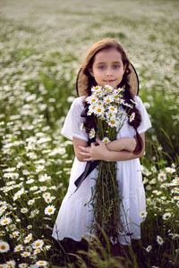 Portrait girl child in a white dress stands on a camomile field in a hat. bouquet of flowers