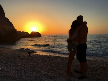 Side view of couple embracing while standing on shore at beach against sky during sunset