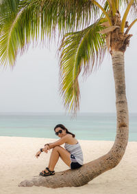 Woman sitting on palm tree at beach against sky