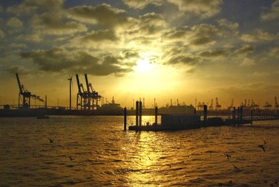 Silhouette cranes at harbor against sky during sunset