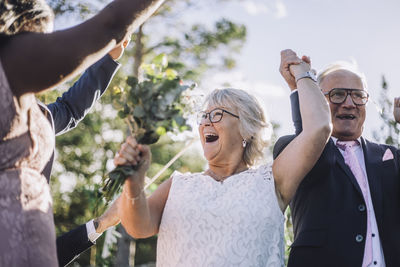 Cheerful newlywed senior couple holding hands while dancing with family on wedding day