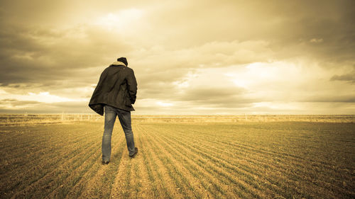 Full length rear view of man walking on plough field against cloudy sky