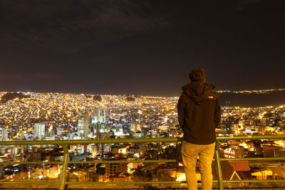 Rear view of man looking at illuminated cityscape against sky at night