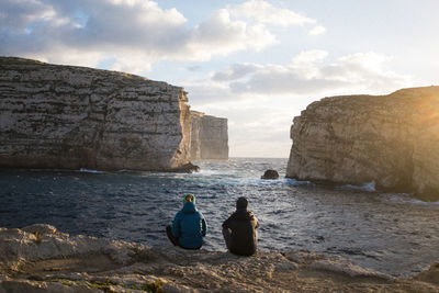 Rear view of men sitting on rock by sea against sky
