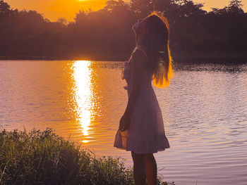 Rear view of woman standing at lakeshore during sunset