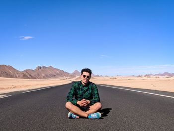 Portrait of handsome young man wearing sunglasses sitting on road against blue sky at desert