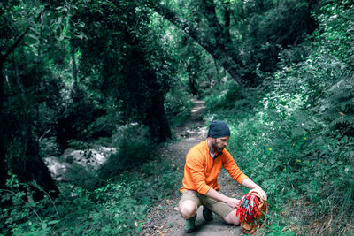 Calm bearded male tourist with backpack sitting on trail in green woods during summer vacation