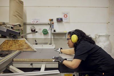 Side view of female carpenter wearing protective eyewear and ear protectors while working in factory