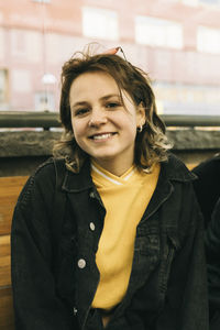 Portrait of smiling young woman wearing denim jacket while sitting on bench