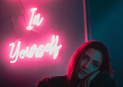 Portrait of young woman sitting against pink neon text on wall