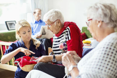 Female caretaker assisting senior women in knitting while man reading book in background at nursing home