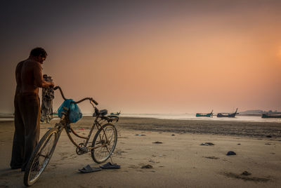 Man on bicycle at beach against sky during sunset