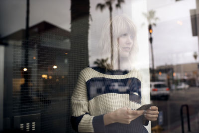 Thoughtful young woman holding smart phone seen through glass wall