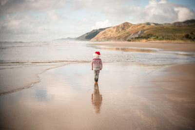 One child wearing a red holiday hat walking on a beach