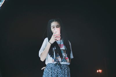 Low angle view of young woman photographing while standing against sky during night