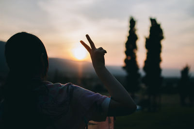 Rear view of silhouette woman showing peace sign against sky during sunset