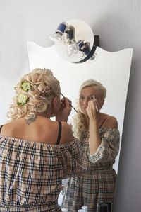 Rear view of woman applying eye make-up while looking in mirror against wall