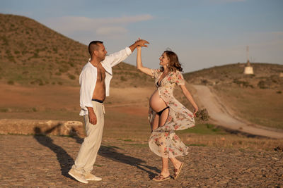 Pregnant woman with dancing man on footpath against sky