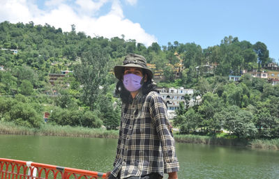 A long haired young man wearing sun hat and face mask, posing by lake with looking at camera.