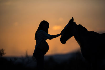 Silhouette pregnant women touching horse against sky during sunset