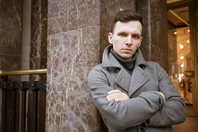 A young man stands outside a shop on the street in a gray coat leaning.