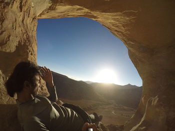 Close-up of man in cave by landscape against sky in sunny day