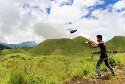 Side view of young man throwing rock while standing against cloudy sky