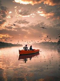 Birds flying over people sailing in lake against sky during sunset