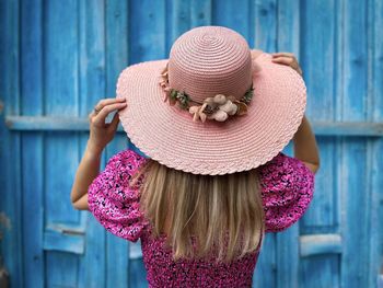 Rear view of blonde woman wearing pink dress and hat facing a blue wooden door