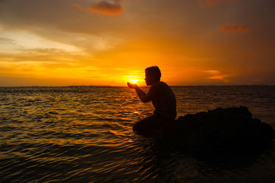 Man sitting on rock at beach against sky during sunset