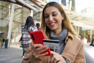 Happy beautiful woman holding her smartphone and enters credit card number on winter season outdoors