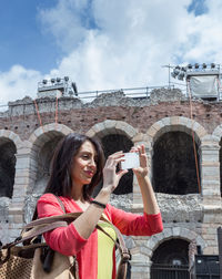 Smiling woman photographing through mobile phone while standing by coliseum