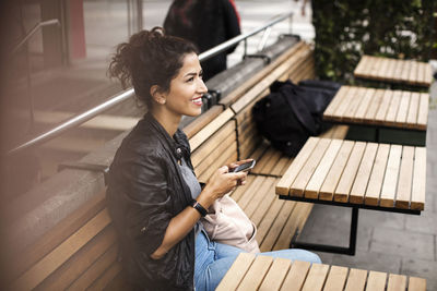 Smiling woman holding mobile phone while sitting on wooden bench at sidewalk cafe