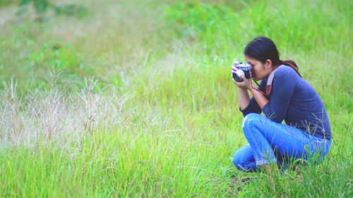 Young woman photographing with camera while crouching on grassy land