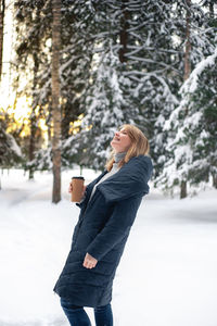 A nice middle-aged blonde woman walks through the winter forest with a cup of hot drink. 